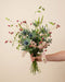 Eine Hand hält den lockeren Wildblumenstrauss Bergluft mit blauen Edeldisteln und rosa Levkojen vor beigem Hintergrund