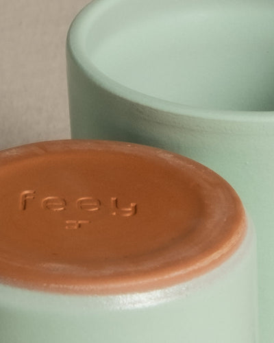 Nahaufnahme des Bodens einer Keramiktasse, auf deren terrakottafarbener Unterseite das Wort „Keramik Baby-Topfset 'Sencillo' (6 Stk.)“ eingeprägt ist. Im Hintergrund ist eine weitere ähnliche Tasse zu sehen, beide mit einer glatten, pastellgrünen Außenseite, perfekt für das Keramik Baby-Topfset 'Sencillo' (6 Stk.).