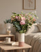 Eine Ballerina Mit Vase, gefüllt mit rosa und weißen Blumen und grünem Laub, steht auf einem Holztisch neben einer Tasse in einem gemütlichen, sanft beleuchteten Schlafzimmer mit einem ordentlich gemachten Bett im Hintergrund.