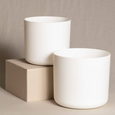 Zwei einfache, weiße, zylindrische Topfsets (18, 16) werden vor einem neutralen Hintergrund präsentiert. Ein Topf steht auf einem quadratischen, beigen Block, während der andere direkt auf der Oberfläche ruht. Beide haben ein glattes, minimalistisches Design.