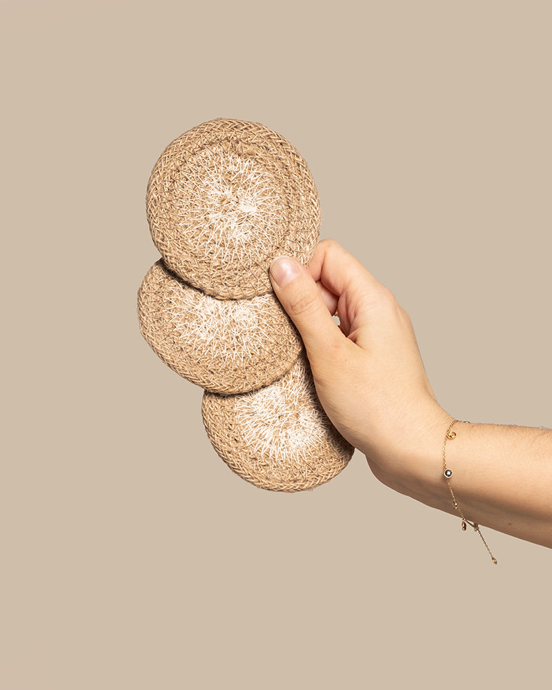 Eine Hand, die drei runde Babypflanzen-Untersetzer aus Jute vor einem beigen Hintergrund zeigt, während das Handgelenk von einem zarten goldenen Armband geschmückt wird. Es ist eine Szene, die detailreiche Handarbeit widerspiegelt, bei der jedes Element sorgfältige Handwerkskunst verkörpert.