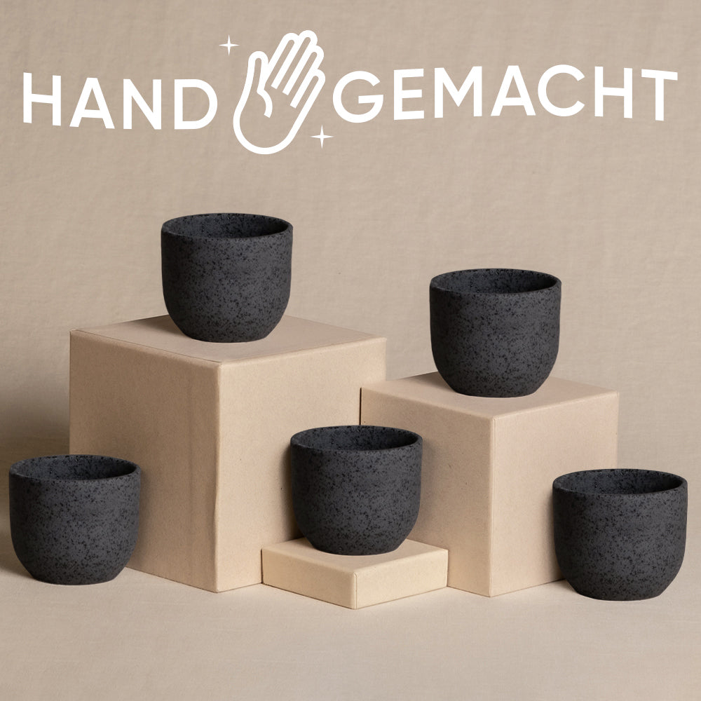 Ein Display mit fünf schwarzen Babypflanzen-5er-Topfsets Keramik ist auf beigen Sockeln vor einem passenden Hintergrund angeordnet und verleiht dem Ambiente eine natürliche Note. Über den Tassen ist der Text „HAND GEMACHT“ zusammen mit einem abgebildeten Handsymbol deutlich zu sehen.