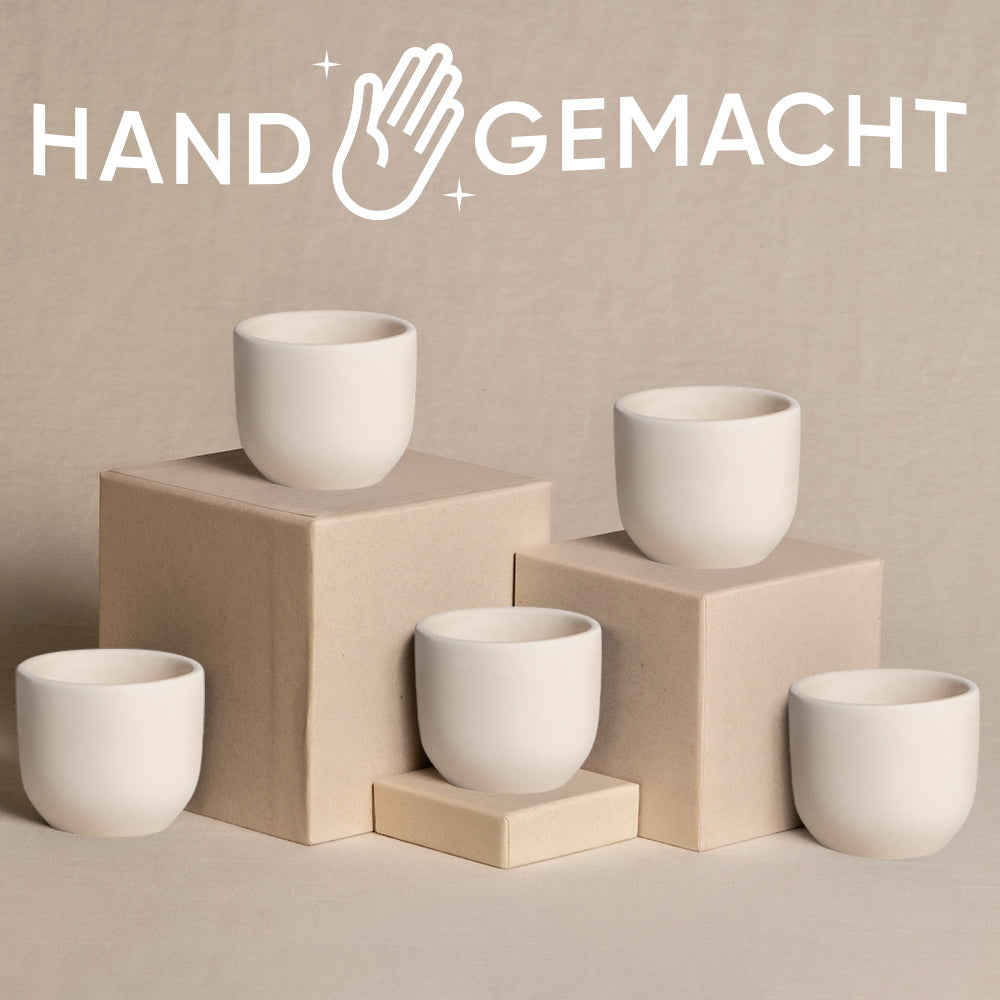 Fünf weiße, minimalistische Keramiktassen werden auf hellbraunen Boxen auf neutralem Hintergrund präsentiert. Darüber steht das Wort „HANDGEMACHT“ mit einem Handsymbol, was darauf hinweist, dass sie handgefertigt sind. Diese eleganten Stücke, die dem zarten Babypflanzen-5er-Topfset Keramik ähneln, verleihen jeder Umgebung eine natürliche Note.