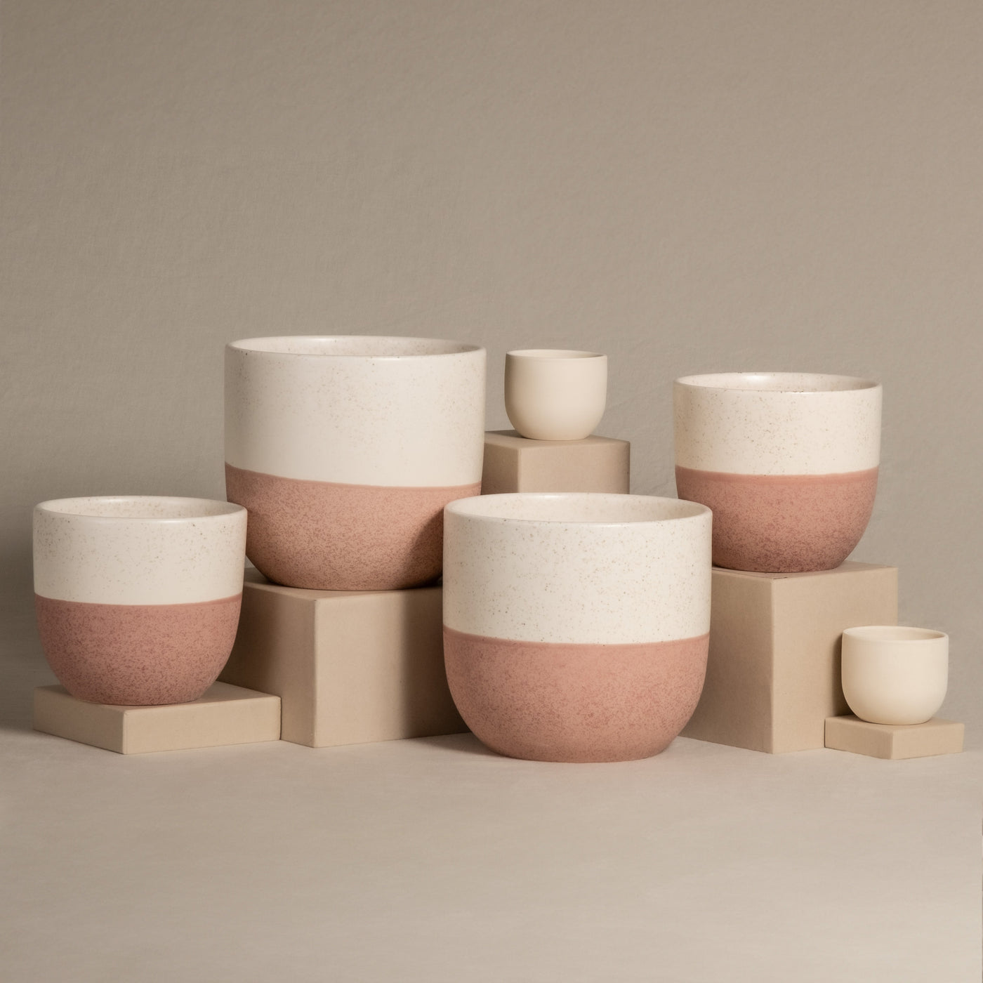 Set aus weißen und rosa Keramik-Topfsets „Variado“ (2 × 18, 2 × 14, 2 × 7) in verschiedenen Größen, dargestellt auf beigen Blöcken vor neutralem Hintergrund. Die Pflanzgefäße sind zweifarbig gestaltet, wobei der obere Teil weiß und der untere Teil strukturiert rosa ist, perfekt für die Präsentation Ihrer Zimmerpflanzen.