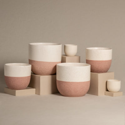 Set aus weißen und rosa Keramik-Topfsets „Variado“ (2 × 18, 2 × 14, 2 × 7) in verschiedenen Größen, dargestellt auf beigen Blöcken vor neutralem Hintergrund. Die Pflanzgefäße sind zweifarbig gestaltet, wobei der obere Teil weiß und der untere Teil strukturiert rosa ist, perfekt für die Präsentation Ihrer Zimmerpflanzen.