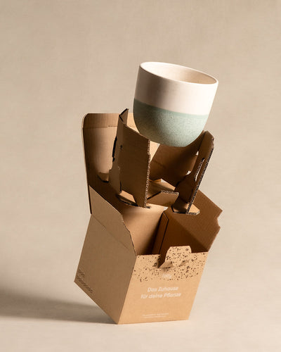 Ein Keramiktopf mit weißem Deckel und mintgrünem Boden wird von einer aufwendigen Kartonverpackung getragen. Die Verpackung ist teilweise geöffnet und gibt den Blick auf das Keramik-Topfset „Direito“ (4 × 14) im Inneren frei. Die Schachtel hat ein minimalistisches Design und einen deutschen Text.