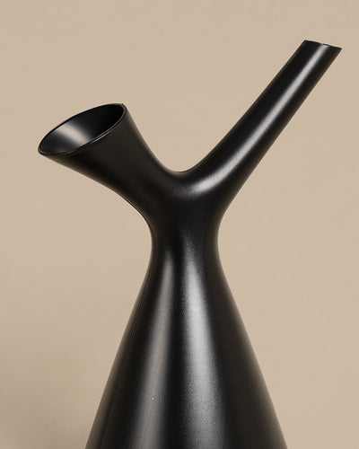 Eine schlanke, schwarze, moderne Plunge Gießkanne mit minimalistischem Design, einem konischen Körper und zwei in entgegengesetzte Richtungen weisenden Ausgüssen, ideal zum Gießen von Pflanzen, vor einem beigen Hintergrund.