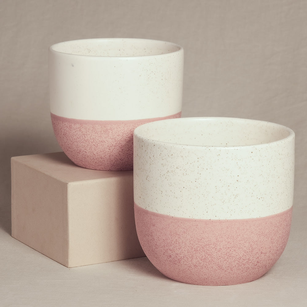 Zwei moderne Keramik-Topfsets 'Variado' (18, 16) mit minimalistischem Design stehen vor einem neutralen Hintergrund. Jeder Topf hat eine weiße obere Hälfte und eine rosa untere Hälfte mit gesprenkelter Textur. Ein Topf steht leicht erhöht auf einem beigen rechteckigen Block, perfekt für die Präsentation von Zimmerpflanzen.