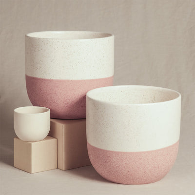 Drei handgefertigte Keramik-Pflanzgefäße unterschiedlicher Größe, jeweils mit einer weißen oberen Hälfte und einer rosa unteren Hälfte, werden auf beigen Blöcken präsentiert. Das kleinste Pflanzgefäß ist schlicht weiß und auf einem Block platziert, während die beiden größeren Keramik-Topfsets 'Variado' (2×16, 7) daneben stehen. Der Hintergrund ist ein neutraler Stoff, perfekt für Zimmerpflanzen.