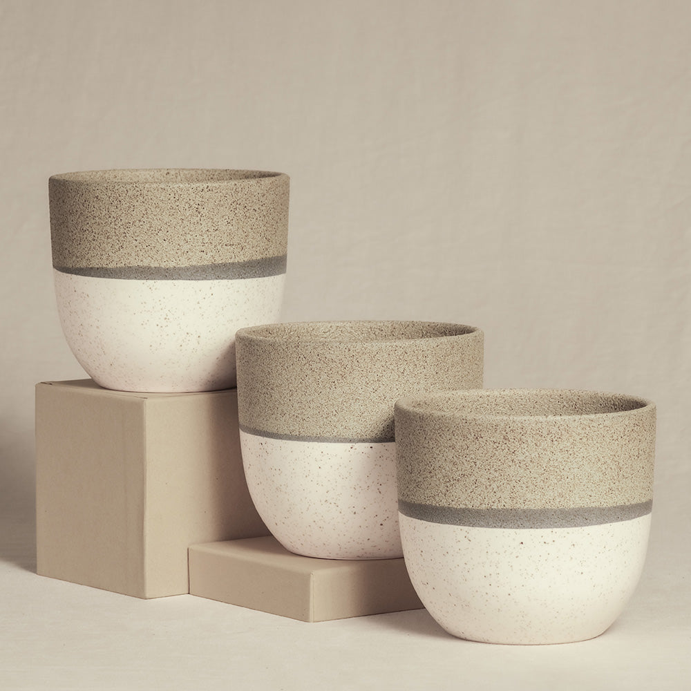 Es werden drei Keramik-Pflanzgefäße in minimalistischem Design gezeigt. Die von Hand hergestellten Pflanzgefäße variieren in der Höhe und sind auf beigen Blöcken angeordnet. Jedes Pflanzgefäß hat eine strukturierte, sandfarbene obere Hälfte und eine glatte, weiße untere Hälfte mit einem grauen Band, das die beiden Abschnitte trennt – perfekt für Zimmerpflanzen. Diese sind Teil des Keramik-Topfsets „Variado“ (3 × 14).