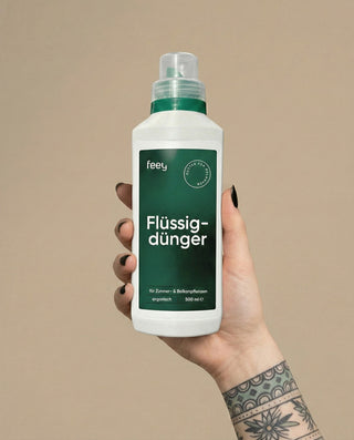 Eine Hand mit schwarzem Nagellack und einer Tätowierung hält eine weiße Flasche mit der Aufschrift "feey Flüssigdünger", ein organischer Flüssigdünger, der für Zimmer- und Balkonpflanzen geeignet ist. Der Hintergrund ist beige.