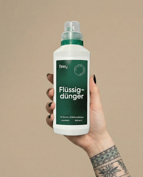 Eine Hand mit schwarzem Nagellack und einer Tätowierung hält eine weiße Flasche mit der Aufschrift "feey Flüssigdünger", ein organischer Flüssigdünger, der für Zimmer- und Balkonpflanzen geeignet ist. Der Hintergrund ist beige.