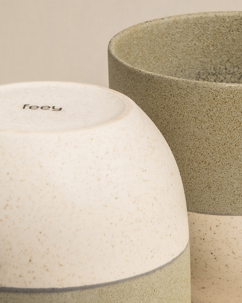 Nahaufnahme von zwei handgefertigten minimalistischen Keramik-Pflanzgefäßen in Erdtönen. Das Pflanzgefäß links ist weiß mit gesprenkeltem Muster und hat das Wort „feey“ auf seinem Boden aufgedruckt. Das etwas größere Pflanzgefäß rechts ist beige mit einer rauen Textur, perfekt für Zimmerpflanzen. Dies ist das Keramik-Topfset „Variado“ (18, 16).
