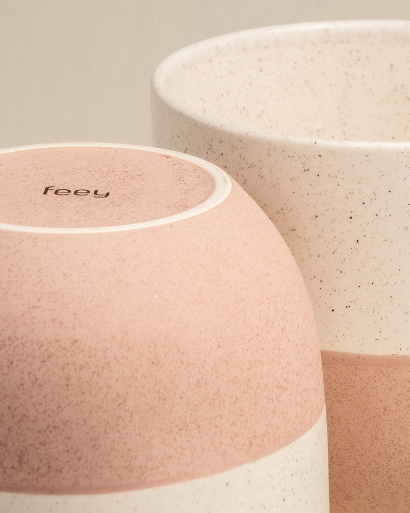 Zwei Keramik-Topfsets 'Variado' (2 × 16, 7) sind abgebildet, eines kopfüber im Vordergrund und das andere aufrecht daneben. Die handgefertigten Tassen haben ein minimalistisches Design mit einem gesprenkelten Muster in Beige- und Hellrosatönen. Auf der Unterseite der Tasse im Vordergrund steht das Wort „feely“.