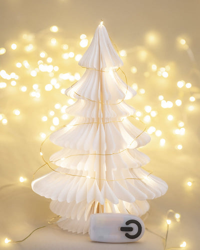 Ein dekorativer Weihnachtsbaum aus weißem Papier ist mit einer Lichterkette geschmückt. Vor dem Baum liegt eine kleine Fernbedienung. Der Hintergrund wird durch eine zusätzliche unscharfe Lichterkette sanft beleuchtet und schafft eine warme und festliche Atmosphäre, die perfekt für die Weihnachtsdekoration ist.