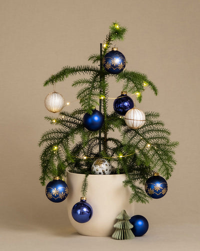 Ein kleiner Norfolk-Tannenbaum im Topf ist mit blauen und weißen Ornamenten dekoriert und mit einer Lichterkette geschmückt. Neben dem Topf steht eine grüne, keramische Miniatur-Christbaumfigur. Der Hintergrund ist beige und bietet einen neutralen Kontrast.