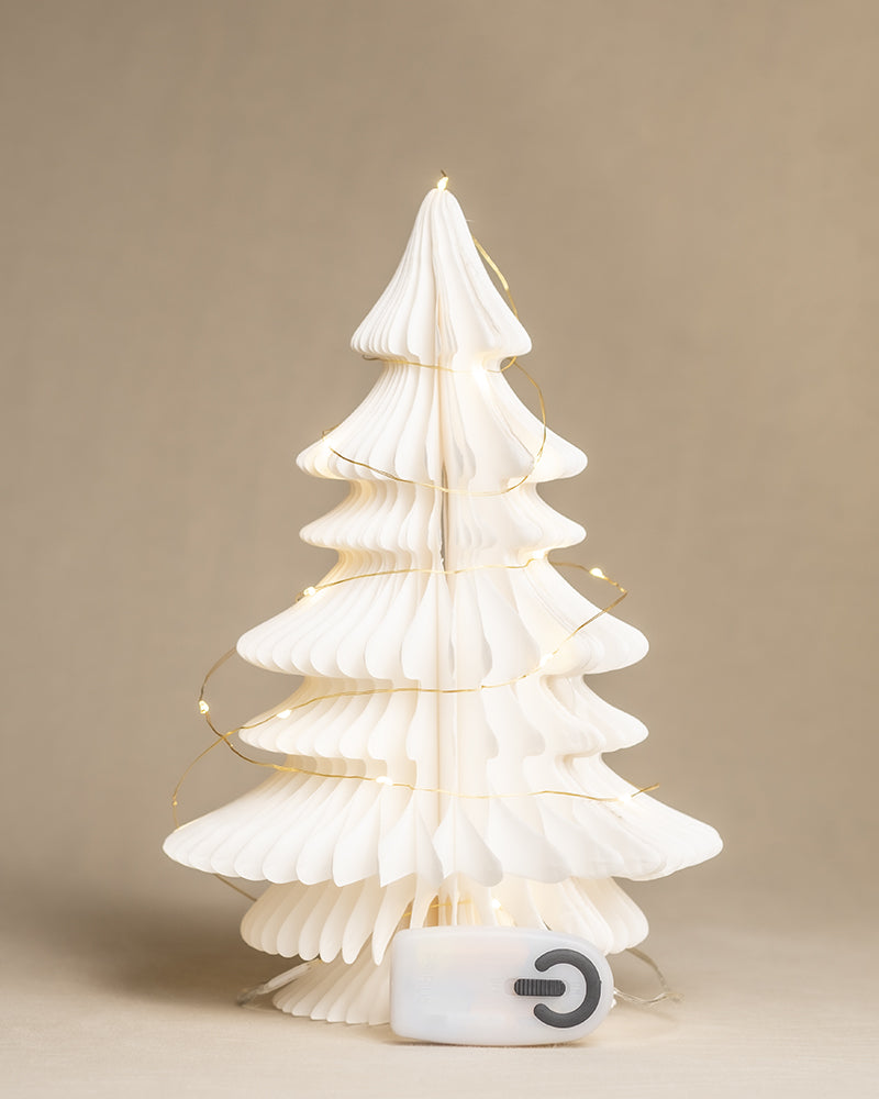 Ein weißer, aus Papier gefalteter Weihnachtsbaum ist mit einer Reihe kleiner, warmer Lichterketten geschmückt. Am Fuß des Baumes sitzt ein kleiner weißer Block mit einem großen Einschaltknopfsymbol. Der Hintergrund ist schlicht beige.