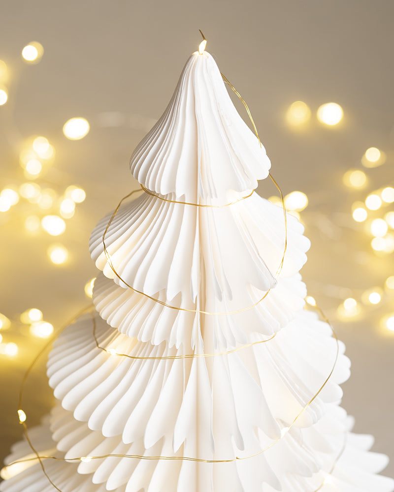 Ein dekorativer Weihnachtsbaum aus weißem Papier, geschmückt mit einer zarten goldenen Lichterkette. Der Hintergrund weist ein sanftes, warmes Leuchten der zusätzlichen verschwommenen Lichterkette auf und schafft eine festliche und gemütliche Atmosphäre, um Ihren Raum weihnachtlich zu dekorieren.