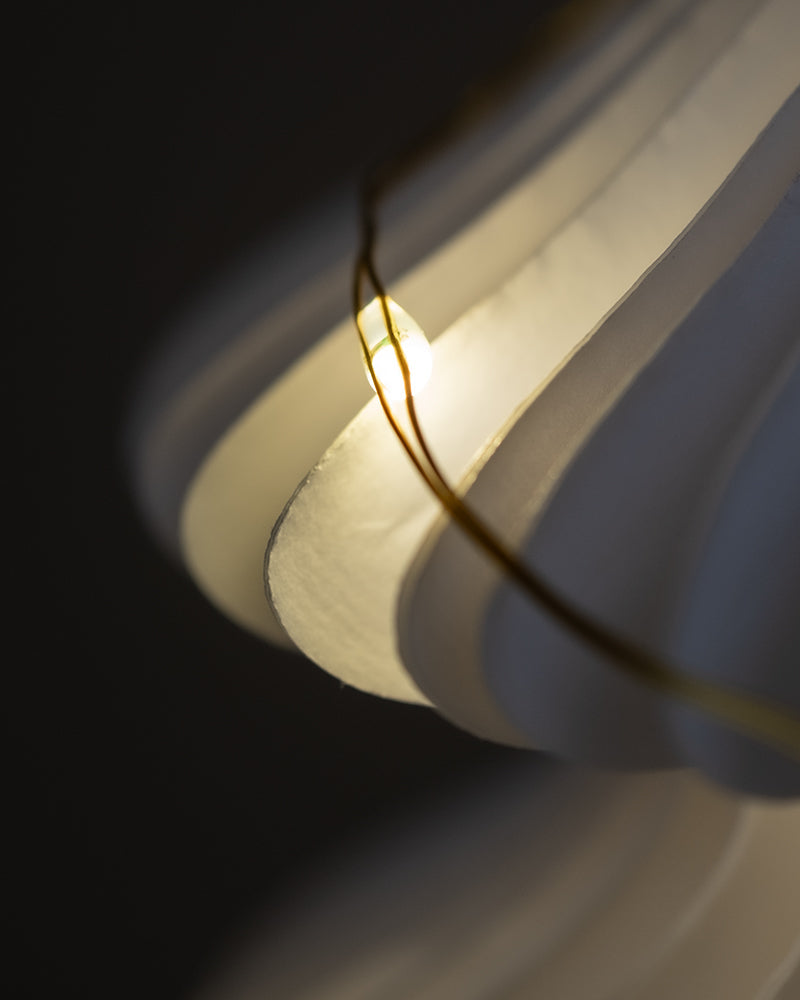 Nahaufnahme einer warm beleuchteten, modernen Pendelleuchte mit geschwungenen, mehrschichtigen weißen Schirmen. In der Nähe der Lichterkette ist ein dünner, goldener Draht zu sehen, der der Komposition ein künstlerisches Element verleiht. Der Hintergrund ist dunkel und betont die Sanftheit des Lichts und des Designs.