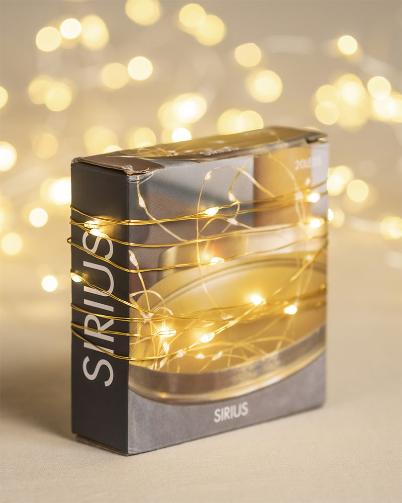 Eine Schachtel mit der Aufschrift „SIRIUS“ ist mit einer Reihe kleiner, warmweißer Lichterketten umwickelt. Der Hintergrund ist mit unscharfen LED-Lämpchen sanft beleuchtet und sorgt so für eine gemütliche, festliche Atmosphäre, die perfekt zum weihnachtlichen Dekorieren ist.